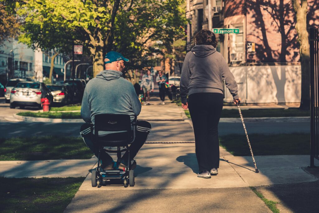 A man in a motorized wheelchair riding on a sidewalk next to a woman walking with a cane.