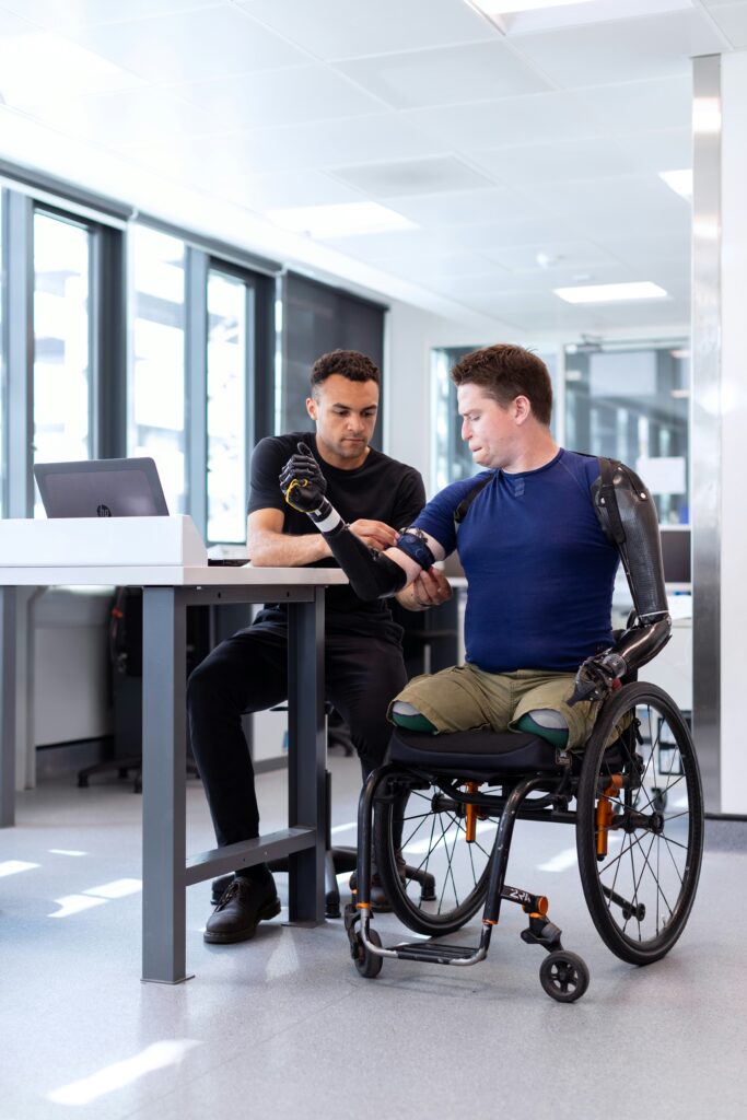 A man with prosthetic arms sits in a wheelchair while another man adjusts his prosthetics.