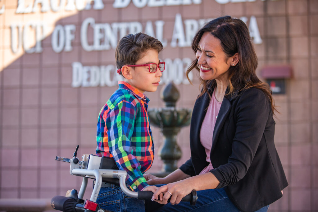 A young boy sitting on a walker while an adult woman kneels down smiling at him.