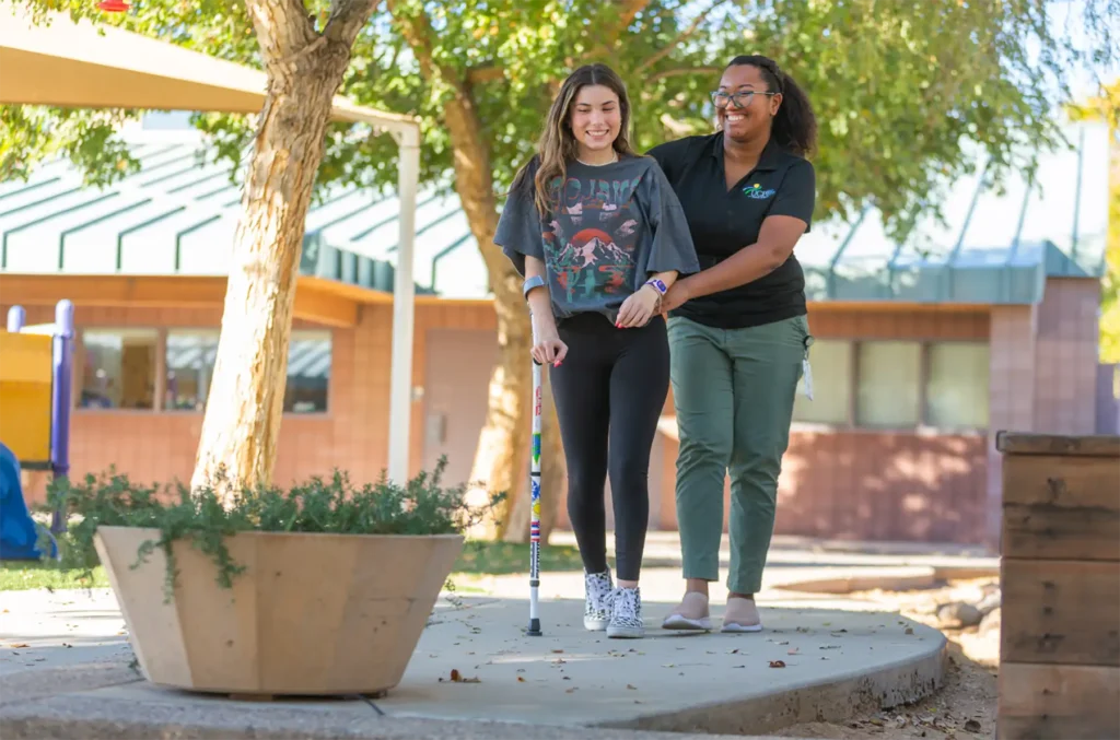 A woman using a crutch walks on a sidewalk while a nurse supports her hips.