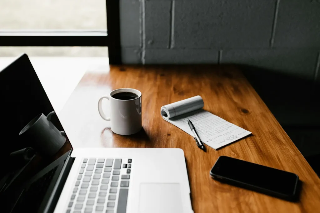 A laptop, coffee mug, notepad, and smartphone on a wooden tabletop