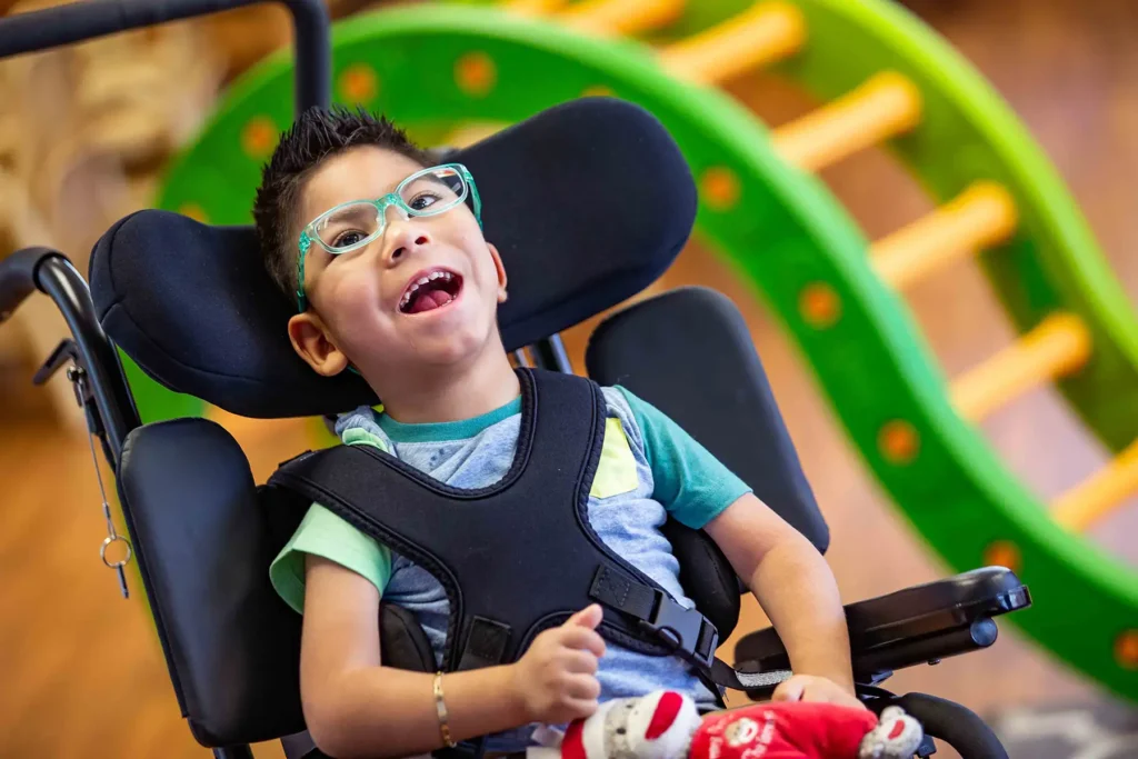 A young boy in a wheelchair in an indoor play area happily smiles.