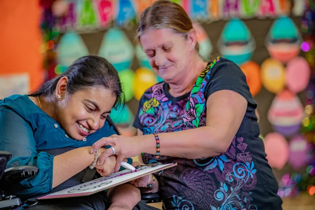 A young woman with cerebral palsy writes on a clipboard with a marker with support from another woman who holds her hand.