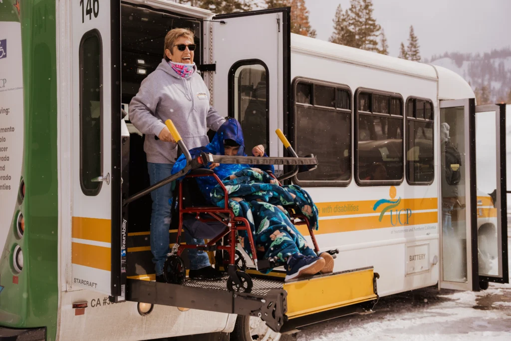 A young man in a wheel chair using a bus ramp while a woman holds the handles of his wheelchair.