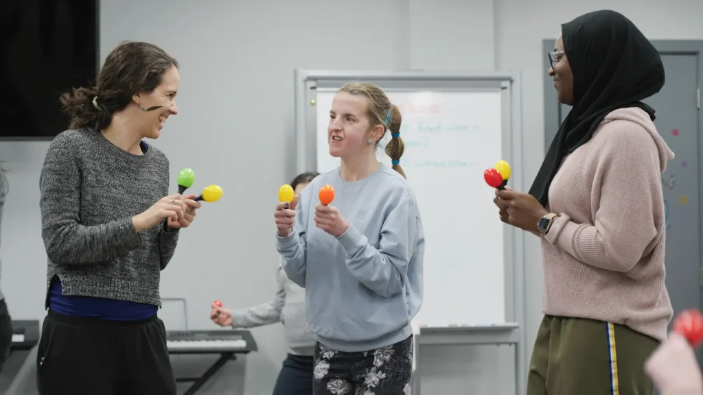 Three women holding maracas stand in a circle making music.