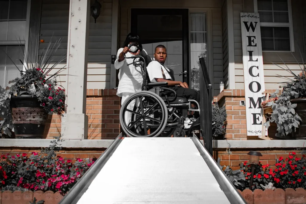 A woman supports a man in a wheelchair above a stair ramp in the exterior of a house.