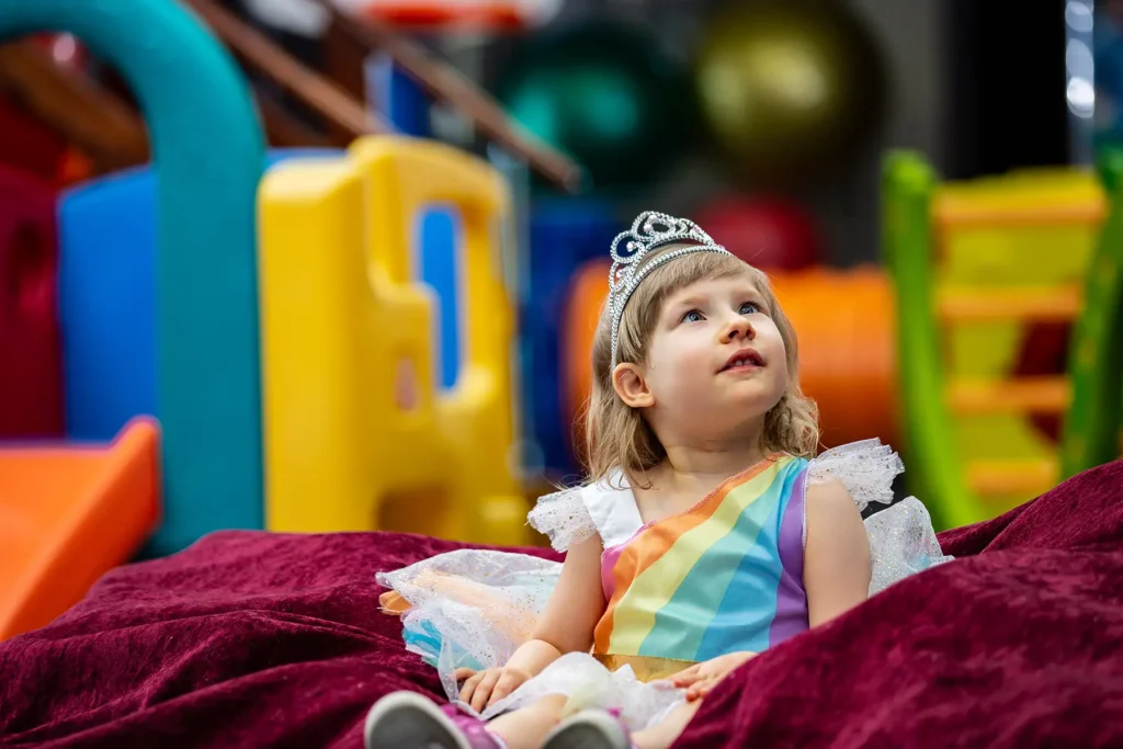 A young girl wearing a princess dress and a tiara smiles in a playroom.