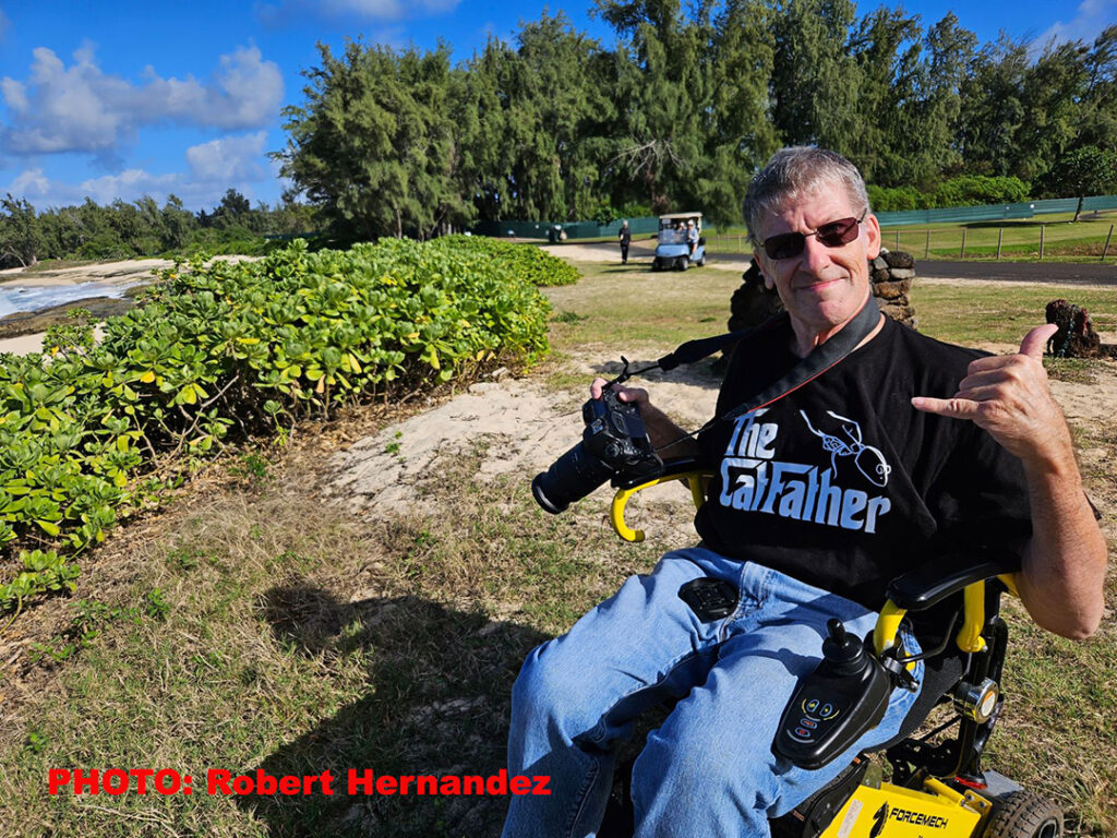 Dr. Stephen Dantzig sits in his wheelchair at a beach in Hawaii. He is holding a camera in one hand and making a shaka gesture in the other.