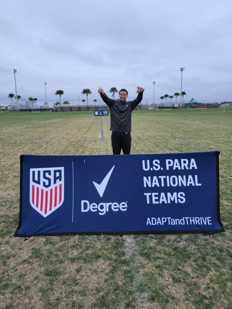 Matt Downing standing on a playing field pointing to a banner for the US Para National Teams