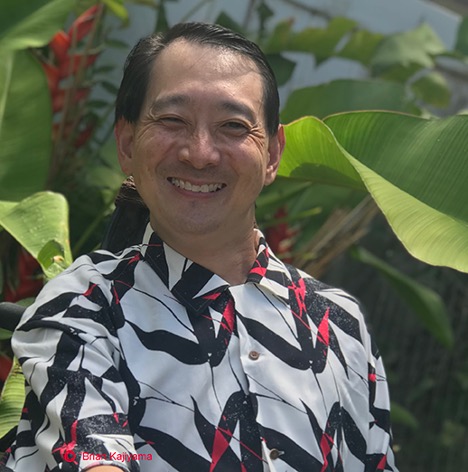 Brian Kajiyama smiling with green foliage in the background.