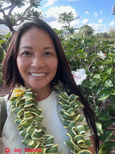 Joy Mahiko wearing a lei and standing with gardens in the background