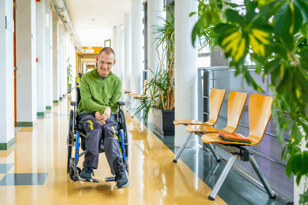 A photo of a man in a wheelchair in a hallway next to a row of chairs.