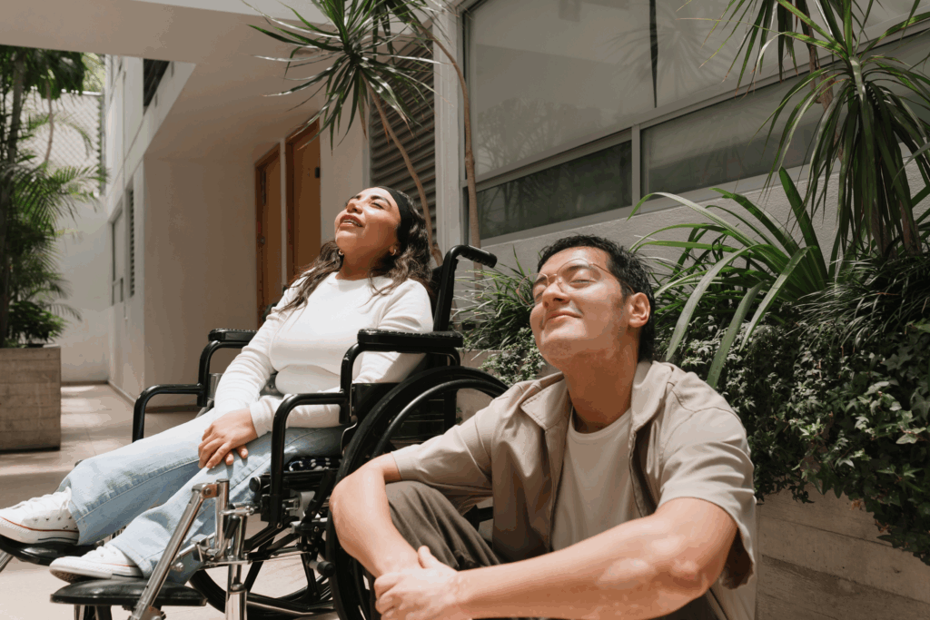 Two people smile with their faced upturned toward the sun. One is sitting on the ground, the other in a wheelchair.