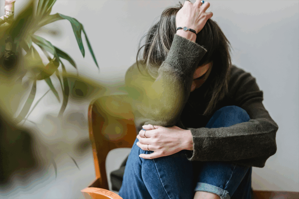 Woman seated in a chair next to plant, she is folded up and hugging herself as if she is sad.
