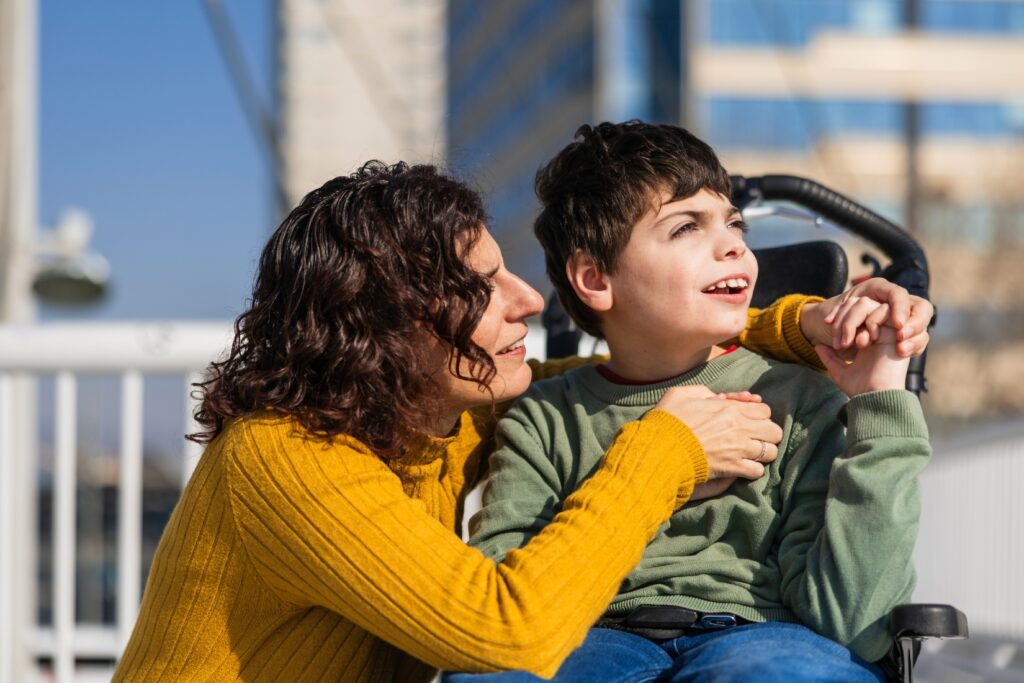 boy with his mother, he is seated in a wheelchair and looking up to the sky while smiling. they are holding hands