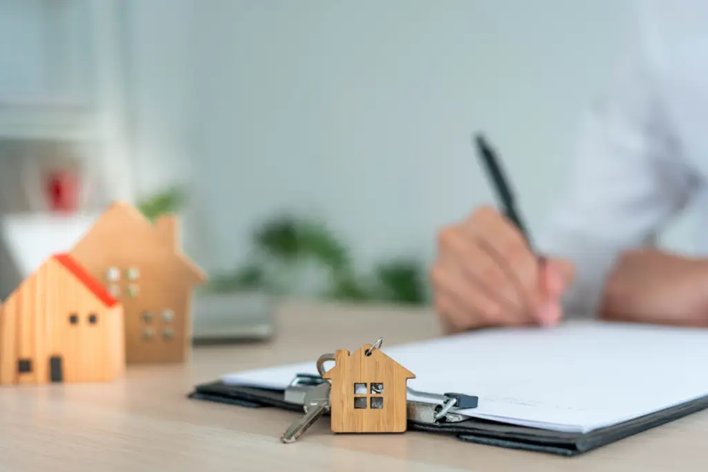 photo of a person signing paperwork with houses on the desk and a house keychain