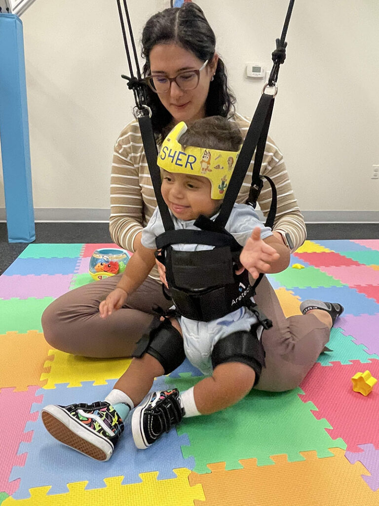 A baby in a harness that is hooked to a machine that helps with gait training. A therapist sits with the child on a colorful mat.